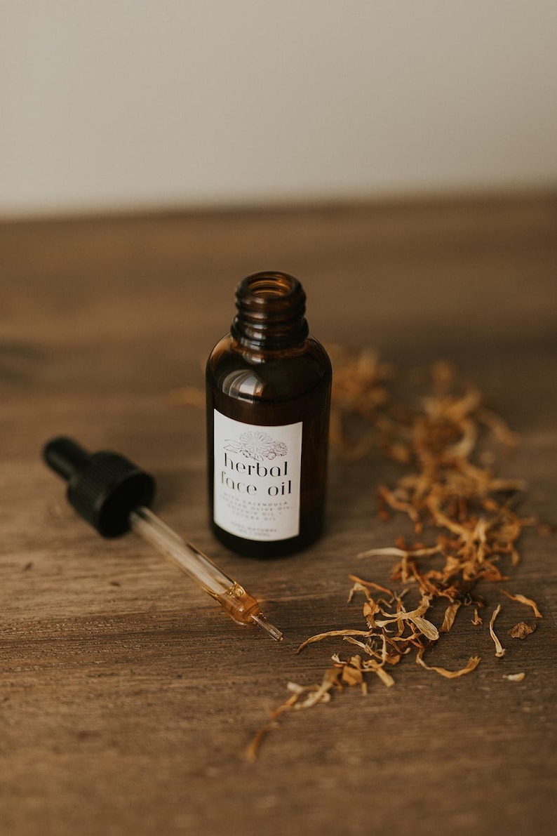 May include: A brown glass bottle of herbal face oil with a dropper, set on a wooden surface. The label reads "herbal face oil". Dried plant matter is scattered around the bottle and dropper.