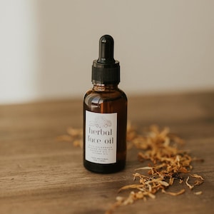 May include: A brown glass bottle of herbal face oil with a black dropper. The label reads "herbal face oil" with additional text. Dried flowers are scattered on the wooden surface. The bottle contains 1 fl oz / 29ml of product.
