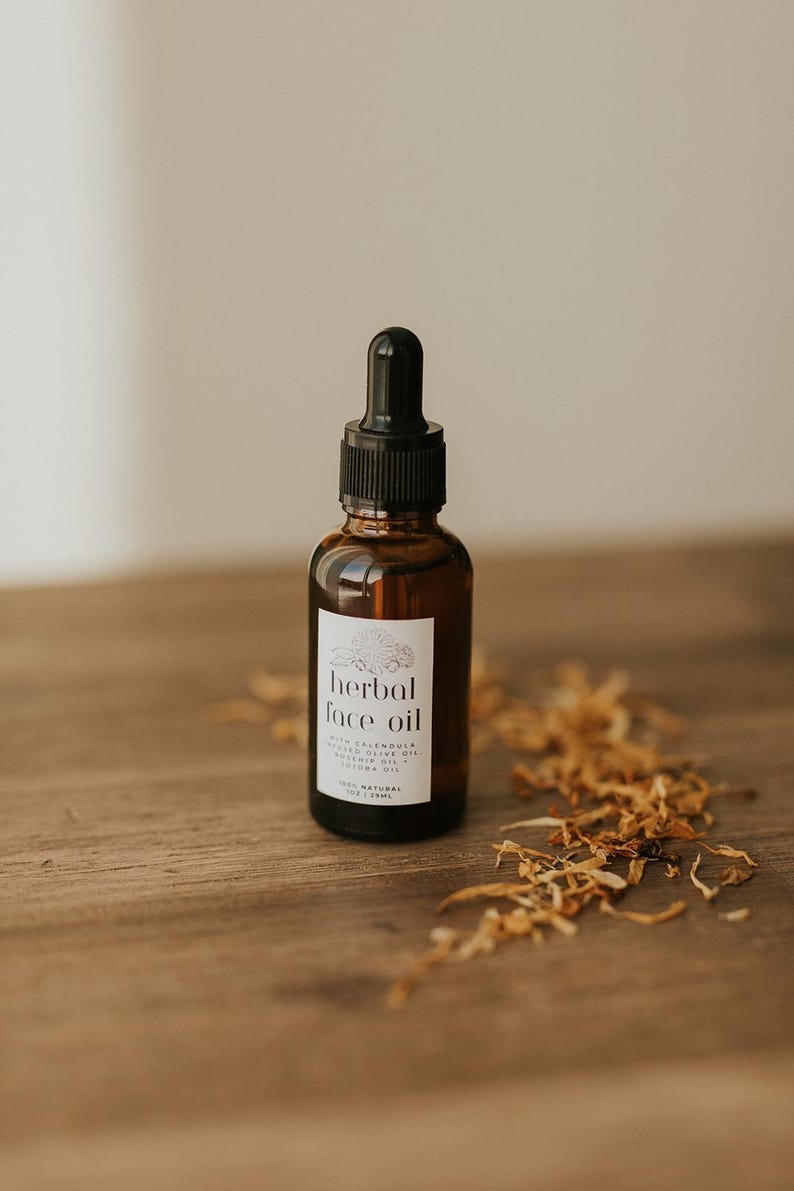 May include: A brown glass bottle of herbal face oil with a black dropper. The label reads "herbal face oil" with additional text. Dried flowers are scattered on the wooden surface. The bottle contains 1 fl oz / 29ml of product.