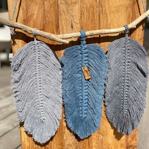 Peut inclure: Trois suspensions murales en macramé en forme de plumes dans des tons de gris et de bleu, avec une branche de bois en haut. La plume du milieu a une petite étiquette en bois avec le mot "Simply" écrit dessus.
