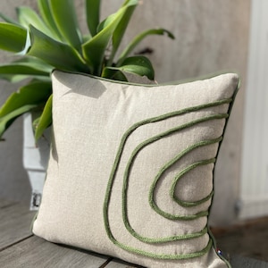 Op de afbeelding: Een beige linnen kussen met een groen touwontwerp. Het kussen heeft een groene rand en ligt op een houten oppervlak.