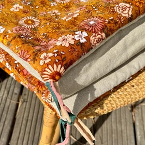 以下が含まれることがあります： 薄い茶色の背景に白とピンクの花が咲いた花柄のクッションが積み重ねられています。クッションはカラフルな布の紐で結ばれており、籐製の椅子の上に置かれています。