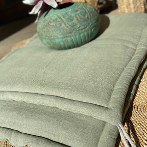 May include: A sage green cushion with a tied string closure. The cushion is made of a soft, textured fabric and has a simple, minimalist design.