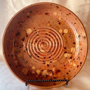 May include: A round ceramic bowl with a speckled terracotta finish, featuring a prominent spiral pattern embossed into the center. The surface displays a mix of warm orange and brown tones with darker flecks and lighter patches throughout the glaze.
