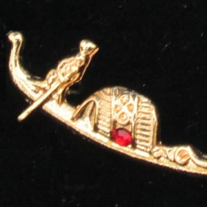 May include: Gold-toned pin featuring an Egyptian-themed design. The pin depicts a stylized boat with a bird figure, a pharaoh's head, and a red gemstone. The pin is likely made of metal and has a decorative, ornate design.