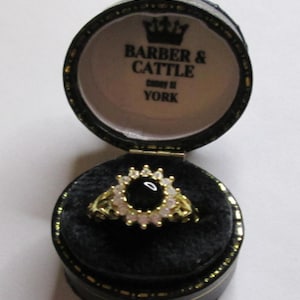 May include: A gold ring with a black center stone and surrounding white stones, displayed in an open black jewelry box. The box has gold trim and the text "BARBER & CATTLE Coney St YORK" is visible inside the lid.