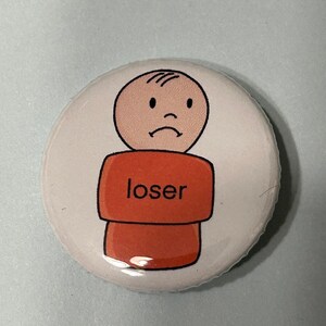 Puede incluir: Un botón blanco con un dibujo animado de una persona triste con la palabra "loser" impresa en su camiseta.