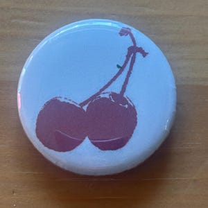 May include: A round button with a light blue background features a stylized illustration of two cherries in a deep red color. The cherries are connected by a stem, creating a simple yet eye-catching design.