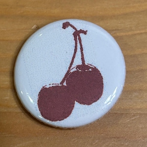 May include: A round white button with a maroon cherry illustration. The cherries are depicted with a stem and two round fruits. The button has a glossy finish and is set against a wooden surface.