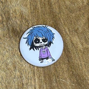 May include: A round, white button featuring a cartoon character with blue hair and a skull face. The character wears a purple sweater with a skull design. The button is a pinback style, suitable for clothing or bags.