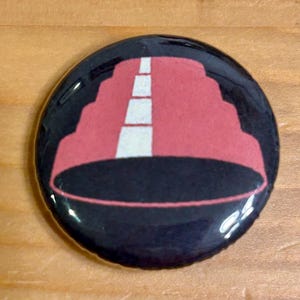 May include: A circular, black button with a glossy surface. It displays a design of a red structure with a white road in the middle. The design is set against a black background.