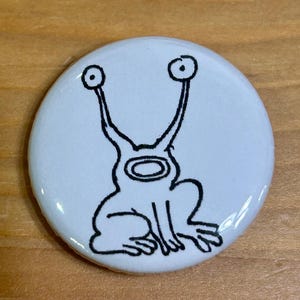 Op de afbeelding: Een witte knoop met een zwarte lijntekening van een cartoonachtig wezen met grote, omhoog wijzende ogen. Het wezen heeft een afgerond lichaam. De knoop ligt op een licht houten oppervlak.