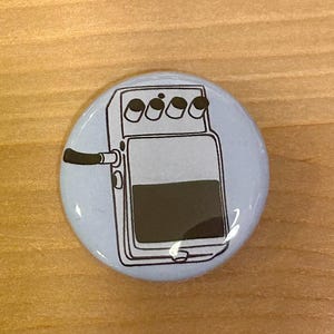 Op de afbeelding: Een ronde, lichtblauwe knoop met een zwart-witte illustratie van een koffiemachine. Het ontwerp omvat een handvat, drie knoppen en een donkerbruine, met koffie gevulde container. De knoop staat op een houten achtergrond.