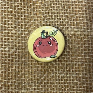 May include: A round, yellow button with a cartoon apple design. The red apple has a sad face and a green worm wearing a hat on top. A green leaf is next to the worm. The button is on a burlap background.