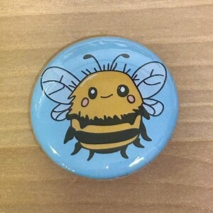 May include: Round button with a cartoon bee design. The bee is yellow and black with a smiling face, set against a light blue background. The button is a fun accessory.