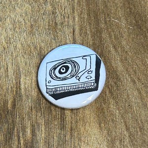 May include: A white button with a black and white illustration of a record player. The button has a glossy finish and is circular in shape. The record player design is a simple line drawing.