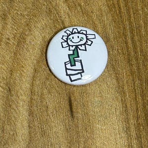 Op de afbeelding: Een witte knoop met een handgetekende afbeelding van een lachende bloem in een pot. De bloem heeft een gele gezicht, groene stengel en staat in een zwart-witte pot. De knoop ligt op een houten oppervlak.