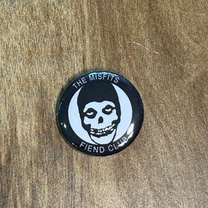 以下が含まれることがあります： Misfits Fiend Clubのロゴが入った、丸い白黒のボタン。デザインは、黒い髪型をした白いドクロが白い三日月を背景にしています。「THE MISFITS」と「FIEND CLUB」の文字が縁に印刷されています。