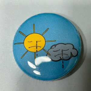 May include: A blue button featuring a yellow sun and a grey cloud, both with sad faces and crying. The sun is on the left and the cloud is on the right.