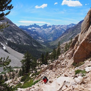 May include: A hiker in a red shirt and backpack walks down a rocky mountain trail. The trail leads to a valley with a view of snow-capped mountains in the distance.