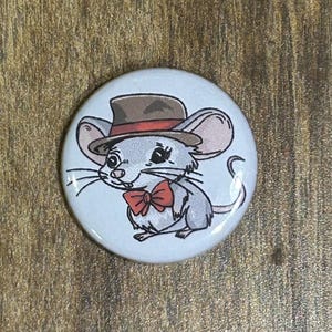 May include: A round, white button featuring a cartoon mouse wearing a brown hat and a red bow tie. The mouse is gray with large ears and long whiskers. The button is set against a wooden background.