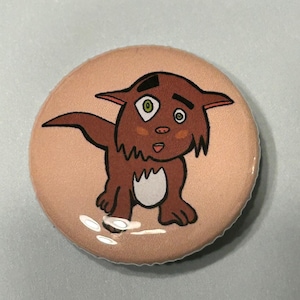 May include: A round button featuring a cartoon illustration of a brown furry creature with green eyes and a pink nose. The creature is standing on a light pink background.
