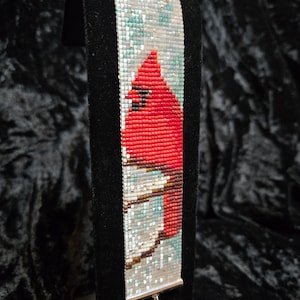 May include: A beaded bracelet featuring a vibrant red cardinal bird design. The bracelet is primarily red, with accents of white, blue, and brown. The bracelet is displayed on a black stand, showcasing the intricate beadwork and color palette.