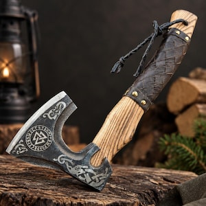 May include: A hand axe with a dark metal head featuring etched Norse knotwork and a Valknut symbol surrounded by runes. The handle is made of light-coloured wood with a dark brown braided leather grip secured by brass rivets and a leather cord lanyard.