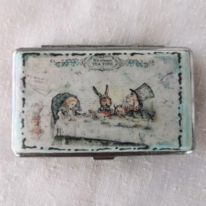 May include: A rectangular, silver-toned metal case with a hinged lid. The lid features a colorful illustration of the Mad Hatter's tea party from Alice in Wonderland, with the text "It's always TEA TIME".
