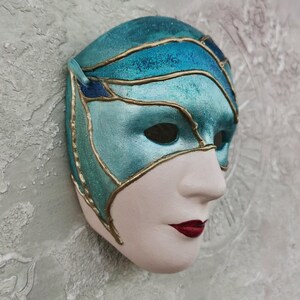 Peut inclure: Un masque de carnaval bleu et or avec un visage blanc et une finition pailletée. Le masque a un design délicat avec des accents dorés et une palette de couleurs bleu et or.