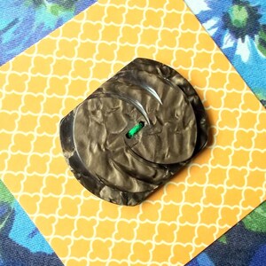 May include: A large, dark grey button with a swirling pattern and a green thread. The button is set on a yellow patterned paper with a blue floral background. The button has two holes for sewing.
