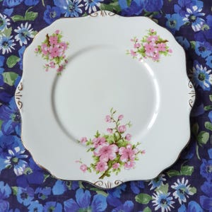 May include: A white square plate with a scalloped edge and gold trim. The plate features pink cherry blossom branches and green leaves. The plate is set against a blue floral patterned background.