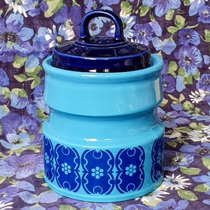 May include: A cylindrical ceramic canister with a glossy light blue finish and a dark blue lid featuring a rounded handle. The base of the canister is decorated with a repeating dark blue geometric pattern that includes small floral motifs.