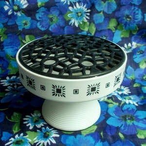 May include: A white pedestal flower arranger with a black geometric top and decorative black square and diamond patterns around the rim. The base is white and ribbed. The background is a blue floral pattern.