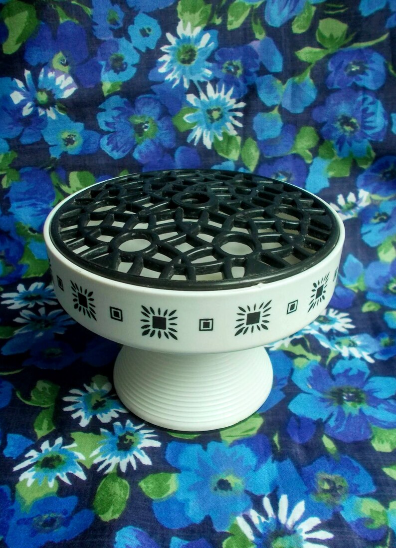 May include: A white pedestal flower arranger with a black geometric top and decorative black square and diamond patterns around the rim. The base is white and ribbed. The background is a blue floral pattern.