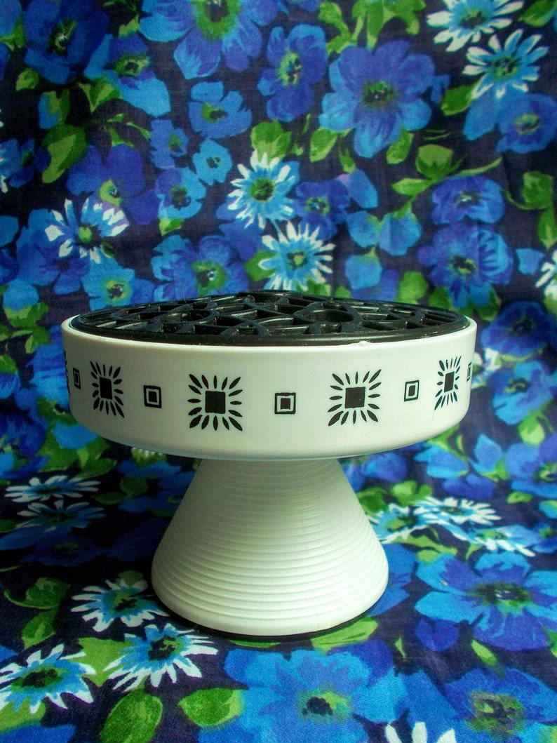May include: A white ceramic flower frog with a black grid top, a decorative band with black geometric designs, and a conical base with horizontal ridges. The background features a blue floral pattern.