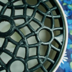 May include: Close-up of a black, ornate, circular object with a lattice design. The object has a white rim and is set against a blue floral background. The design features repeating circular and geometric shapes.