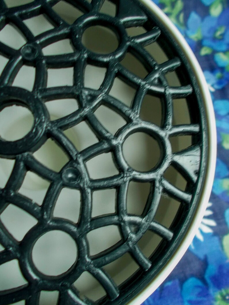 May include: Close-up of a black, ornate, circular object with a lattice design. The object has a white rim and is set against a blue floral background. The design features repeating circular and geometric shapes.