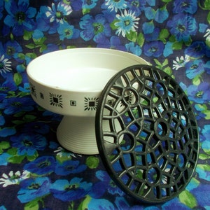 May include: A white pedestal serving dish with a black geometric pattern and a black, openwork trivet. The dish has a wide, shallow bowl and a ribbed base. The trivet is circular with a decorative design. The background is a blue floral pattern.