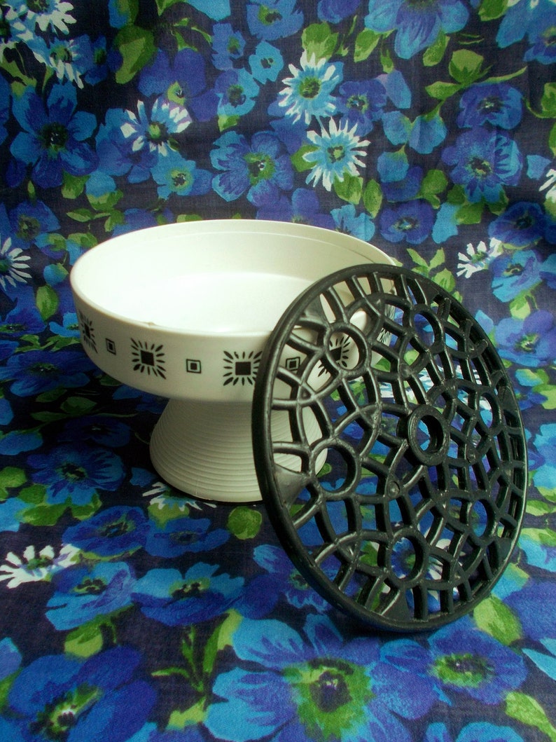 May include: A white pedestal serving dish with a black geometric pattern and a black, openwork trivet. The dish has a wide, shallow bowl and a ribbed base. The trivet is circular with a decorative design. The background is a blue floral pattern.
