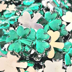 May include: A close-up image of a pile of green and silver shamrock-shaped embellishments. The shamrocks are made of a shiny, reflective material.