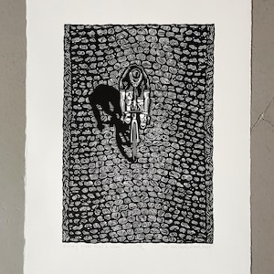 Op de afbeelding: Zwart-witte houtsnede van een fietser die op een geplaveide weg rijdt. De fietser draagt een helm en een trui. De print is getiteld "Keeping the Pedal Turning" en is gesigneerd door de kunstenaar.