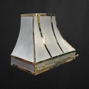 May include: A white metal range hood with a brass trim and a brass handle. The hood has a curved design and is accented with decorative rivets.