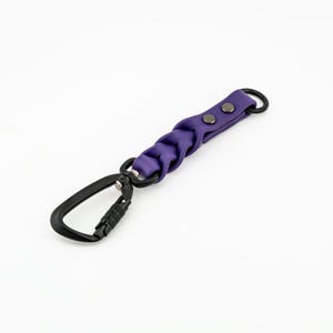 May include: A purple braided leather keychain with a black carabiner clip. The keychain has two silver metal snaps.