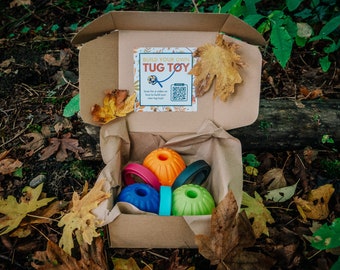 May include: A cardboard box with a brown paper liner contains a build-your-own dog tug toy kit. The kit includes four colorful rubber balls and four rope rings. The box has a label that says "Build Your Own Tug Toy".
