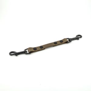 May include: A tan leather dog lead splitter with black metal fittings. The splitter has two black swivel snap hooks and a central section with three black rivets. Designed to split into two leads.