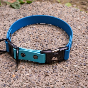 K&ouml;nnte beinhalten: Ein blaues Hundekragen aus Gummi mit einer schwarzen Schnalle und einem silbernen Metallring. Die Schnalle hat ein Dreieckslogo.