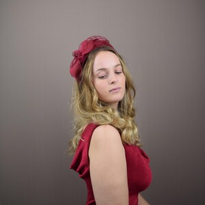 May include: A woman wearing a burgundy red hat with a net veil and a burgundy red sleeveless dress.