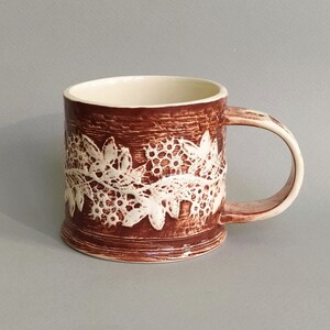 May include: A brown ceramic mug with a white lace design around the middle. The mug has a rounded handle.