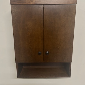 May include: A dark brown wooden wall-mounted cabinet with two doors and a shelf. The cabinet has black knobs on the doors.